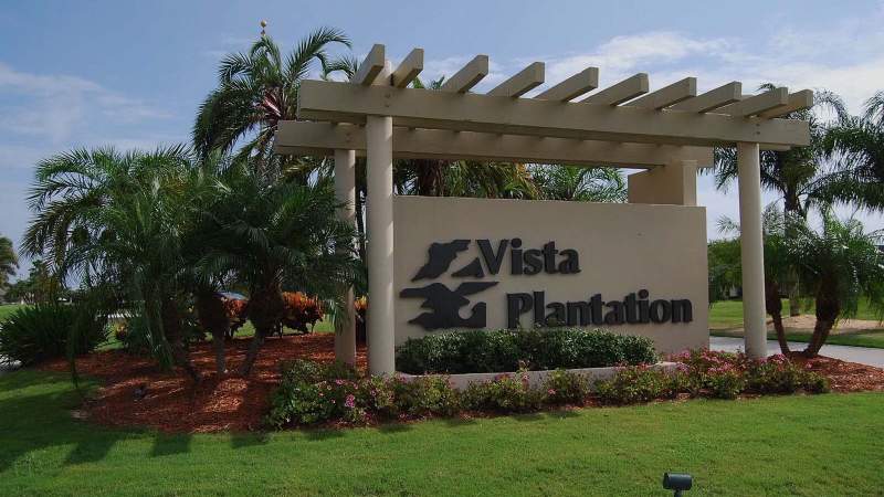 001-White pergola with Vista Plantation sign surrounded by lush greenery and palm trees under a cloudy sky_