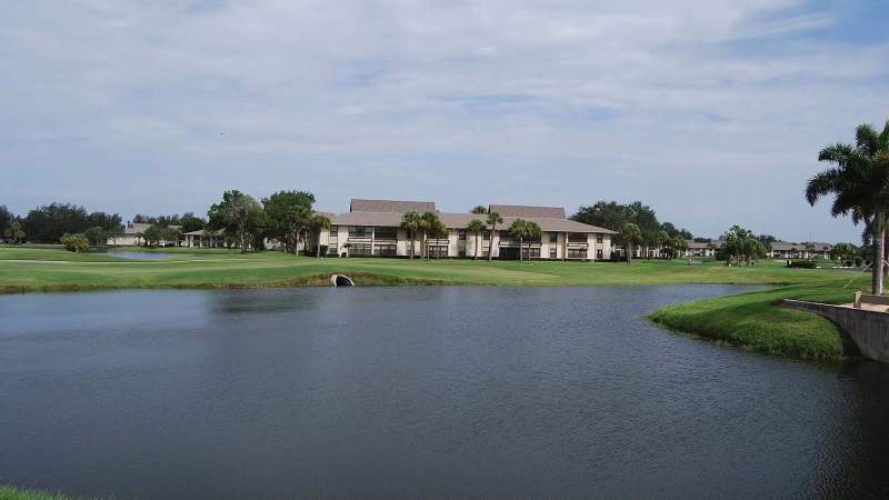 007 - A serene view of a Vista Plantation golf course with a large pond in the foreground, featuring green grass and a multi-unit residential building in the background under a clear sk