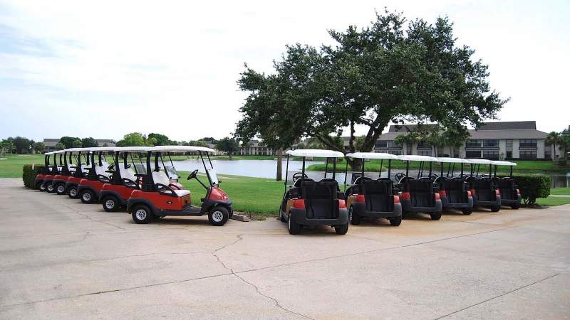 017 - A row of parked golf carts, with one red cart in the front, on a concrete path adjacent to a grassy area and Vista Plantation residential buildings in the background_