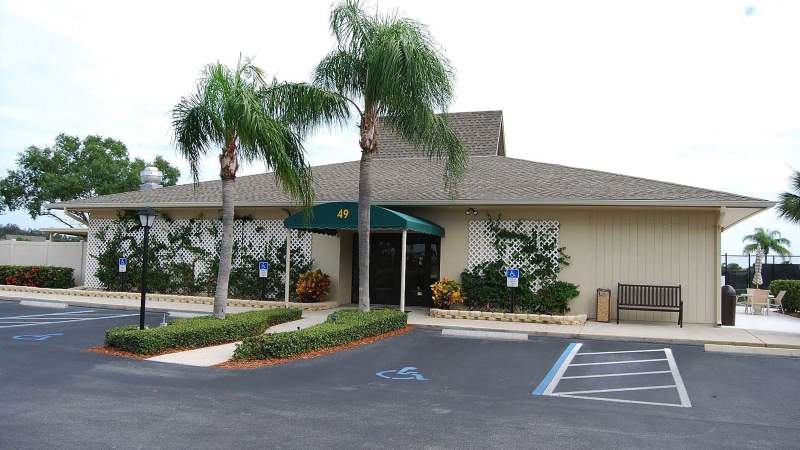 019 - A single-story Vista Plantation building with a beige facade and a green roof, flanked by palm trees, featuring a covered entrance and a parking lot with accessible parking spaces