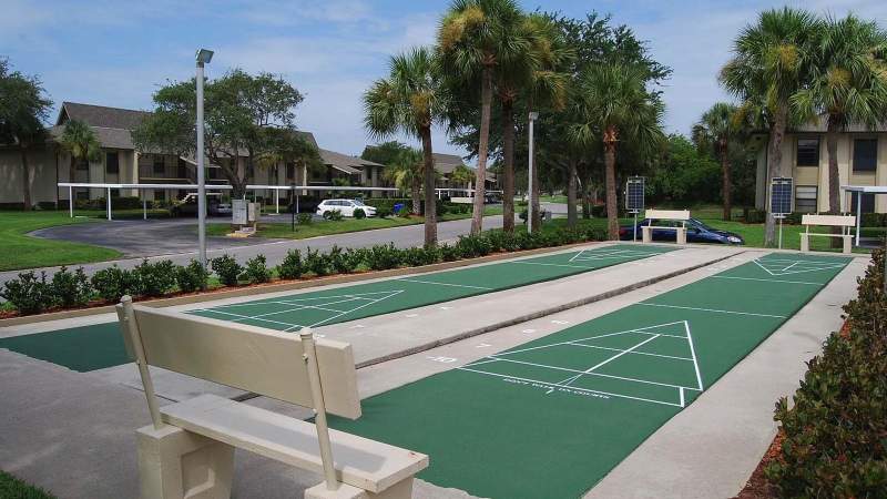 026 - An outdoor shuffleboard court with white boundary lines and numbered sections, surrounded by palm trees and Vista Plantation residential buildings under a cloudy sky_